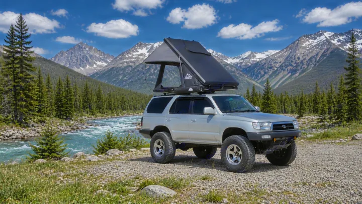 Off-road RV rental: A lifted silver Toyota 4Runner with a rooftop tent deployed, beside a mountain river & snowy peaks.. Toyota 4runner 2001