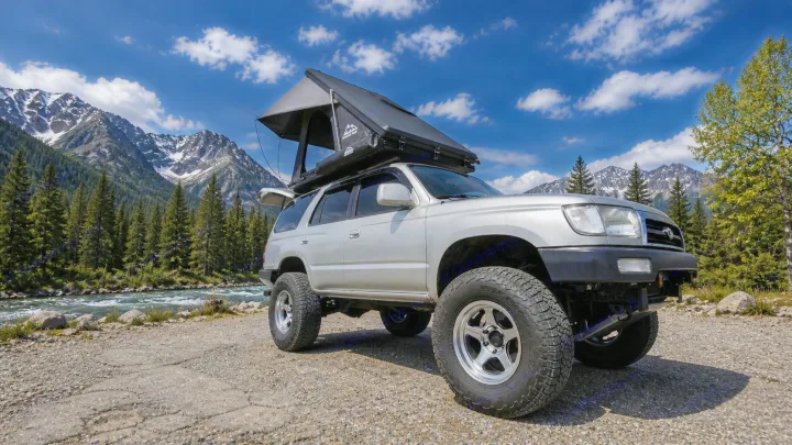 Silver 4Runner with a deployed rooftop tent, perfect for an RV rental adventure. Parked by a river with mountains in the background.. Toyota 4runner 2001
