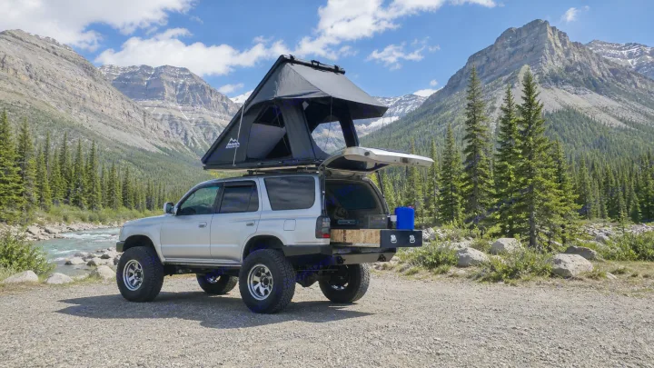 Silver SUV with open rooftop tent & gear, parked by a river with mountains. An ideal off-road camping or rv rental option.. Toyota 4runner 2001