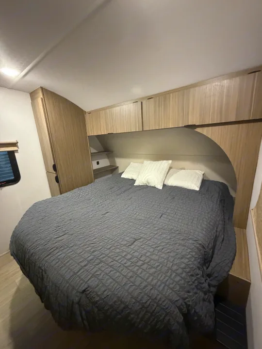 Inside this RV rental, a comfy bedroom offers a large bed with a gray quilted comforter, white pillows, and sleek wood storage cabinets.. Forest River Wildwood X-Lite 2025
