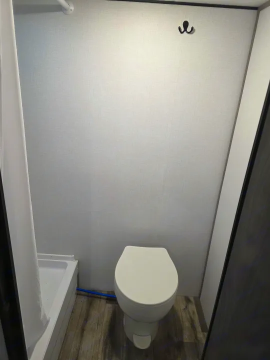 Compact RV rental bathroom with a white toilet, shower, and wall hook. Features light grey textured walls and wood-look flooring.. Coleman Coleman Coleman Lantern Lt 17B 2024