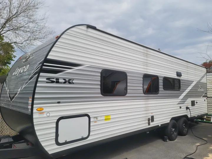 Jayco SLX travel trailer rv rental, parked outdoors. Gray and white side profile with windows and utility connections.. Jayco 260BH 2026