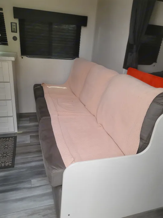 Inside an RV rental, a gray sofa with a pink blanket, white cabinet, and window with black blinds create a cozy living space.. Jayco 260BH 2026