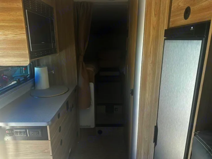 RV rental interior featuring a compact kitchenette with microwave, counter, paper towels, and fridge, leading to a bed area.. Winnebago EKKO 2026