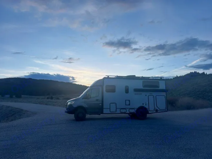 Adventure-ready rv rental parked on a gravel road at dawn/dusk, surrounded by hills & trees under a cloudy sky.. Winnebago EKKO 2026
