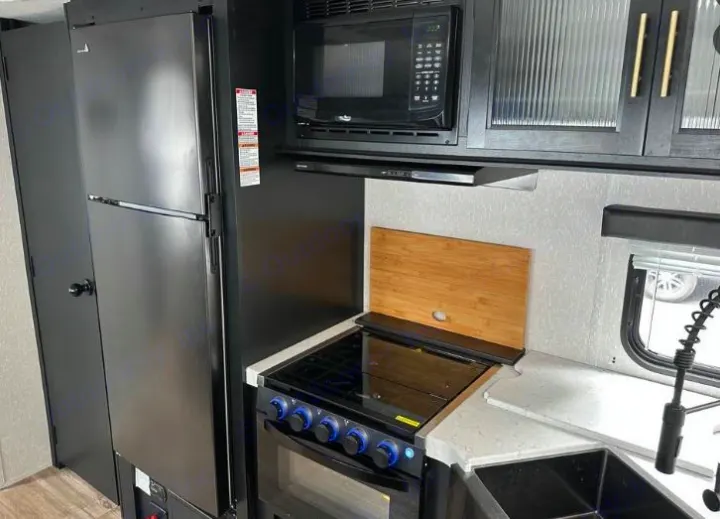 This RV rental kitchen features a compact design with a stainless steel fridge, black microwave, gas stove, and sink area.. Forest River Cherokee Grey Wolf 2022