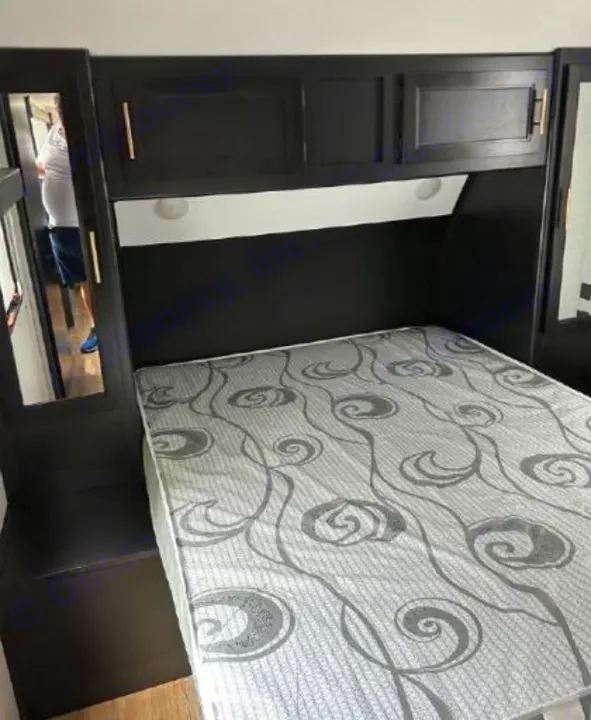 RV rental bedroom featuring a bed with a gray swirl mattress, dark wood overhead cabinets, and built-in side storage.. Forest River Cherokee Grey Wolf 2022