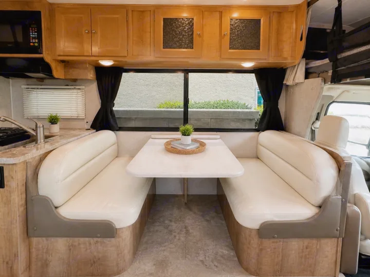 RV rental interior showcasing a cream dinette with a table, and a kitchenette with a sink and wood-toned cabinets.. Forest River Coachmen Leprechaun 2020