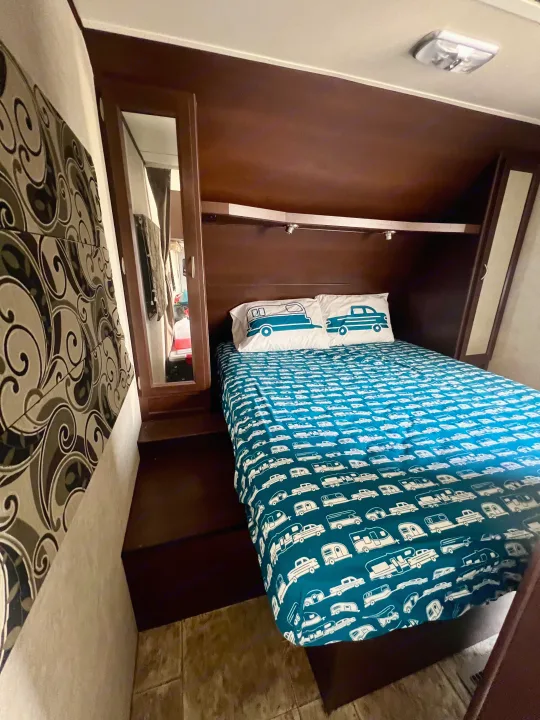 RV rental bedroom features a comfy bed with camper-themed bedding, dark wood cabinets, and mirrored closet doors.. Crossroads Sling Shot 2012