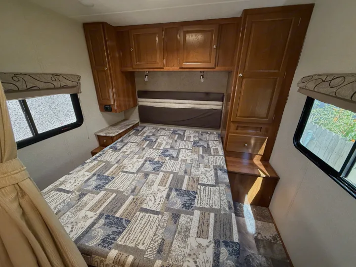 Inside a comfy **rv rental** bedroom: features a patterned bed, wood storage, and natural light from side windows.. Coachmen Leprechaun 2019
