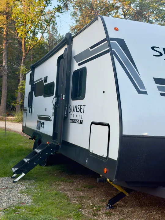 A modern white "Sunset Trail Sport" rv rental with steps down, parked at a wooded campsite.. Crossroads RV Sunset Trail Sport 2025