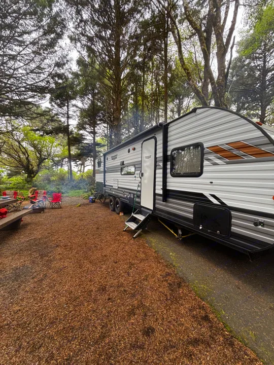 Spacious RV rental at a wooded campsite with a picnic table, fire pit, and chairs. Smoke visibly rises from the fire.. Forest River Wildwood 2020