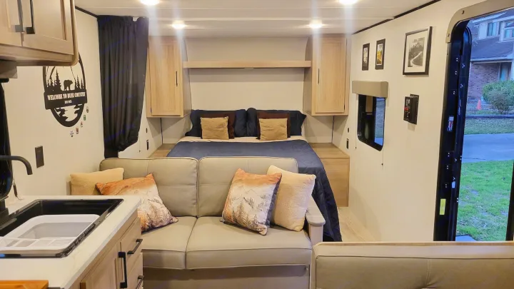 Inside a modern rv rental: a comfy bed, cozy sofa with mountain-themed pillows, and kitchen sink. Bright interior with outside view.. Dutchmen Coleman 2025