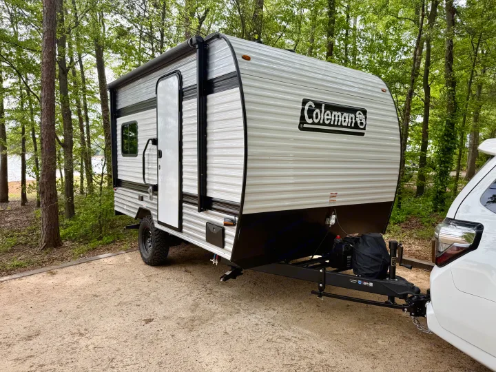 This Coleman travel trailer, an RV rental, is parked at a scenic wooded campsite next to a lake, ready for outdoor adventure.. Keystone Coleman 13T 2026