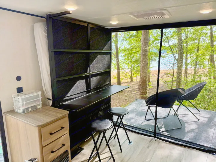 RV rental interior with a versatile workspace & fold-up bed, offering a panoramic view of trees and water from an attached deck.. Keystone Coleman 13T 2026