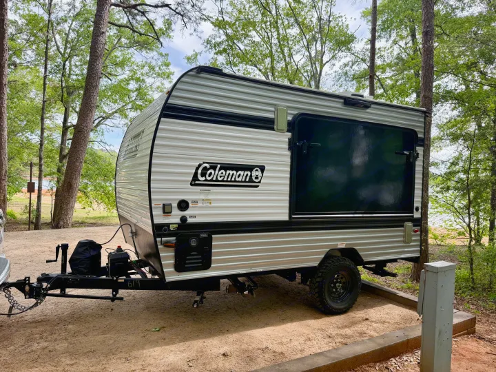 This Coleman travel trailer RV rental is parked on a dirt campsite among trees, with a lake visible in the background.. Keystone Coleman 13T 2026