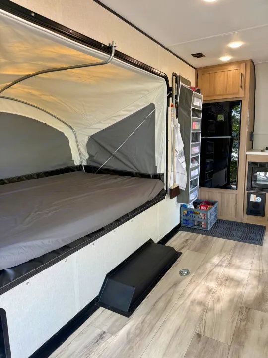 Pop-up RV rental interior: large fold-out bed, kitchenette with fridge, and clever hanging storage. Ready for adventure!. Keystone Coleman 13T 2026