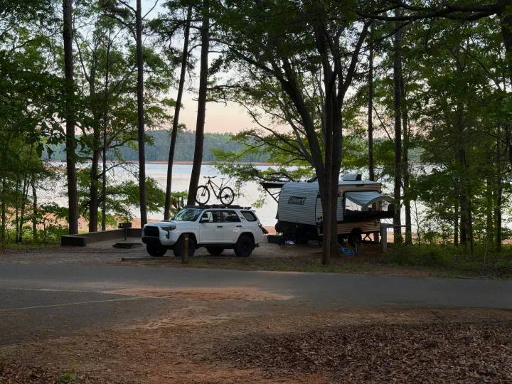 An RV rental and white SUV with a bike on top parked at a scenic lakeside campsite among tall trees.. Keystone Coleman 13T 2026