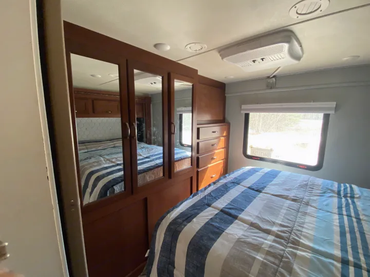 Cozy RV rental bedroom featuring a striped bed, mirrored wardrobe, dresser, window, and ceiling AC unit for a comfortable stay.. Thor Freedom Elite 2016