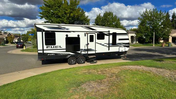 A white 'FUEL' travel trailer rv rental is parked on a suburban street with a flat tire. Notice the residential setting and cloudy sky.. Heartland 265 2021