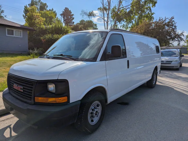 A white GMC cargo van is parked on a sunny street, ideal for an RV rental conversion. Another vehicle is parked behind it.. Gmc Savanna 2500 EXT 2016