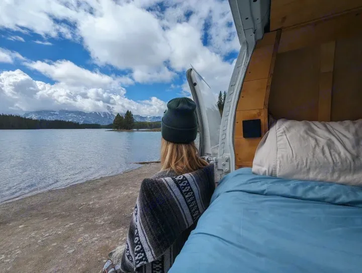 From a cozy RV rental, a person in a beanie and blanket enjoys a serene mountain lake view from the back of their van.. Gmc Savanna 2500 2015