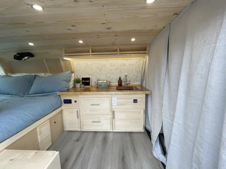 Inside a cozy rv rental van conversion: a comfortable bed, compact kitchen with sink, and light wood paneling throughout.. Gmc Savanna 2500 2015