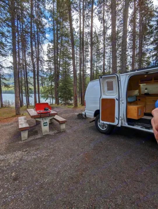 RV rental van at scenic campsite #42 by a lake, surrounded by pine trees, with a picnic table & stove ready for use.. Gmc Savanna 2500 2015