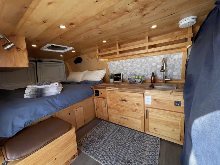 Inviting RV rental interior features a cozy bed, wooden kitchenette with sink, tile backsplash, and ample storage, all in a compact van.. Gmc Savanna 2500 2016