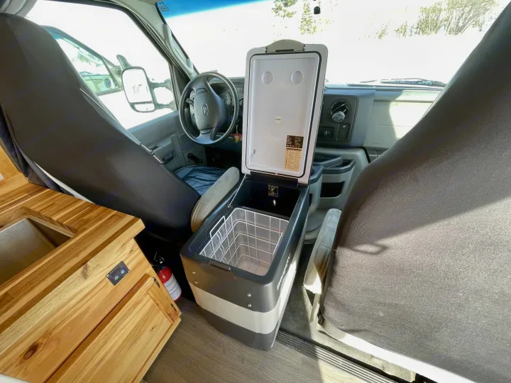 RV rental interior: an open portable fridge between front seats, with a steering wheel and wooden kitchenette featuring a sink.. Gmc Savanna 2500 2016