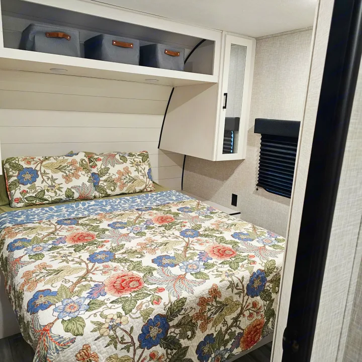 Comfortable RV rental bedroom featuring a bed with a vibrant floral quilt, matching pillows, overhead storage, and a window.. Jayco SLX 2025