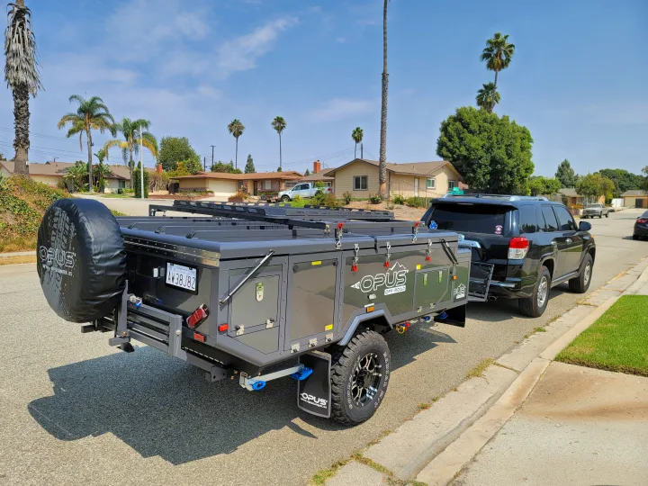 A dark gray Opus off-road camper trailer, ideal for an RV rental, is hitched to a black SUV on a sunny residential street.. Air Opus OP4 2021