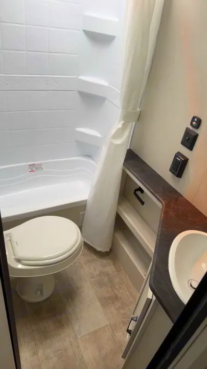 This compact RV rental bathroom features a white toilet, tub/shower with curtain, and a vanity with a sink, all on a light wood-look floor.. Puma Palomino 2024