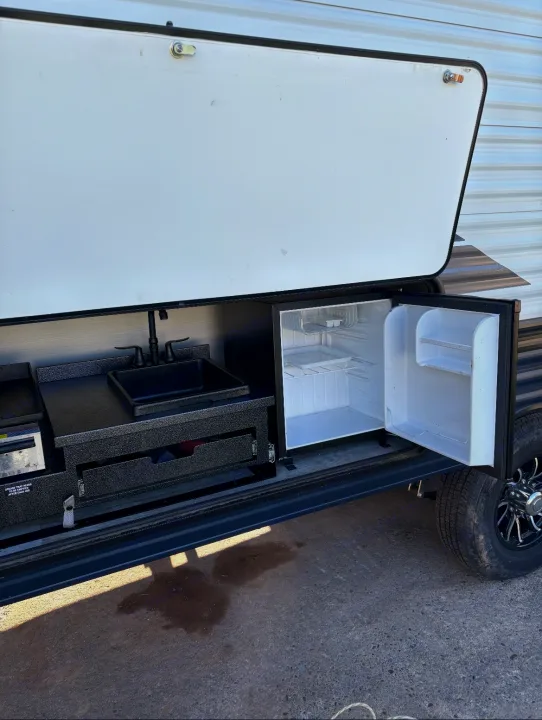 This RV rental's open panel reveals an outdoor kitchen with a sink, faucet, mini-fridge, and cooktop for camping.. Puma Palomino 2024