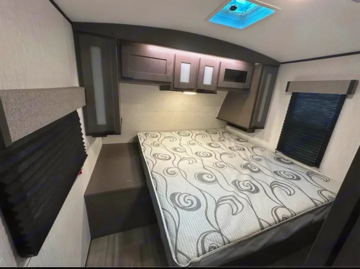 RV rental bedroom with a patterned bed, dark overhead cabinets, and blackout blinds. This compact interior is ready for travel.. Cruiser RV MPG 2022