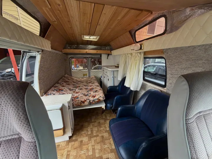 View of a cozy RV rental interior with a wooden ceiling, comfy bed, blue seats, and a compact kitchenette area.. Chevrolet G20 1987