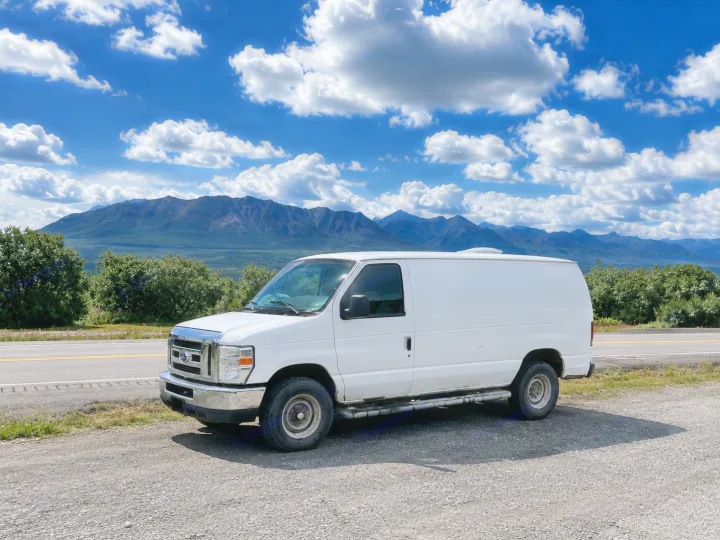 White van, an ideal rv rental, parked roadside with majestic mountains and a vibrant blue sky. Ready for your next road trip adventure.. Ford Econolline 250s 2013