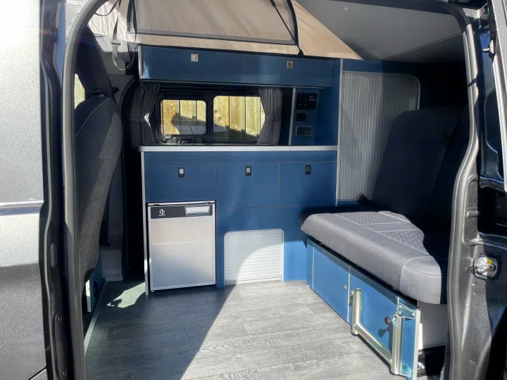 Modern blue interior of an RV rental campervan with a pop-top, fridge, and bench seating, seen from the open side door.. Ford Transit Custom 2+2 berth Pop Top, Automatic 2026