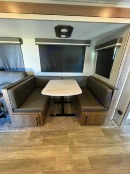 Cozy RV rental dinette with dark grey U-shaped booth seating, a white table, and window blinds. Perfect for meals on the go.. Grand Design Imagine 2022