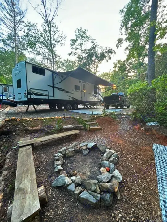 RV rental parked at a woodsy campsite with an extended awning, a fire pit, and rustic wooden benches.. Keystone RV Bullet Premier Ultra Lite 2022
