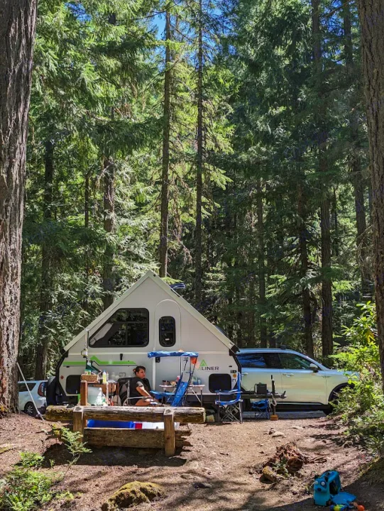 Relaxing at a forest campsite with an A-frame RV rental and a car parked nearby. A person sits enjoying the outdoor setting.. A Liner A Liner Popup Trailer 2022