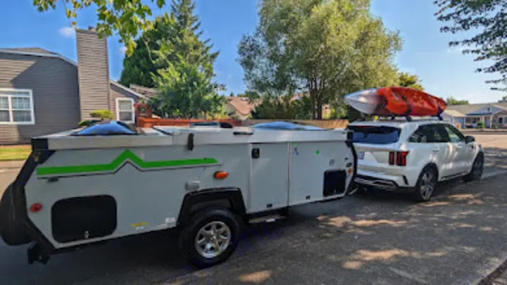 White SUV towing an A-frame pop-up camper with a kayak on top. Ready for your rv rental adventure!. A Liner A Liner Popup Trailer 2022