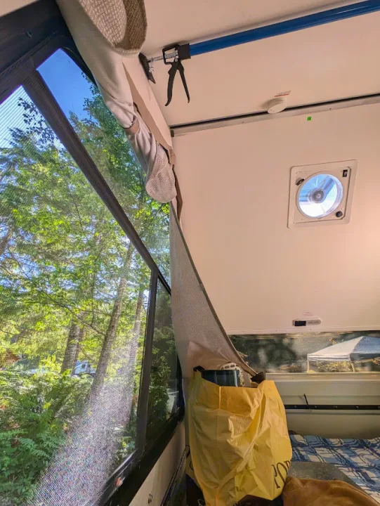Inside an RV rental: A large window overlooks a lush forest campground. Visible are a ceiling fan, curtain, and a blue support pole.. A Liner A Liner Popup Trailer 2022