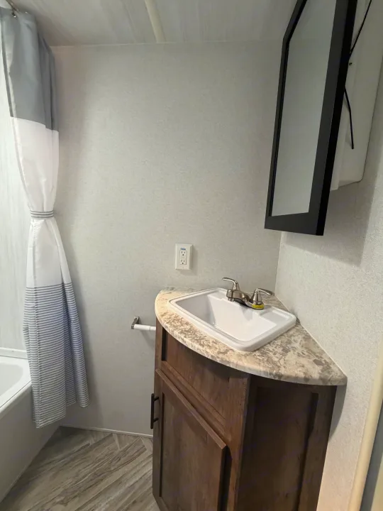 A compact RV rental bathroom with a corner sink, wood vanity, mirror, and a white and grey striped shower.. Forest River Salem Select 2021