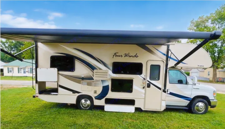 This beige "Four Winds" rv rental features an extended awning and open storage compartments, parked on grass.. Thor Four Winds 2018