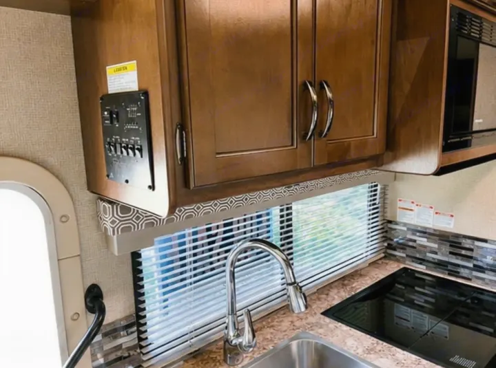 RV rental kitchen featuring wood cabinets, sink, stovetop, and a control panel. A window with blinds offers natural light.. Thor Four Winds 2018