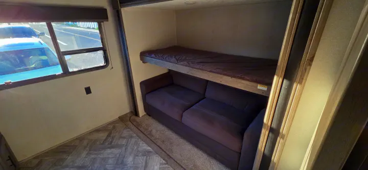 This RV rental interior features a cozy bunk bed above a dark sofa, beside a window with sunlight, and a patterned floor.. Keystone RV Springdale 2021