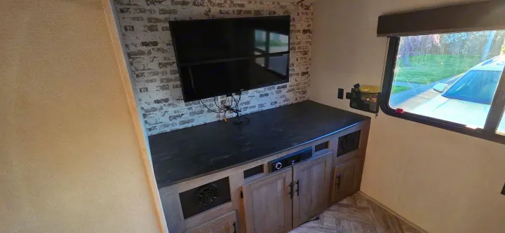 Modern rv rental entertainment area: TV on faux brick, media console with cabinets, and a window overlooking a car.. Keystone RV Springdale 2021