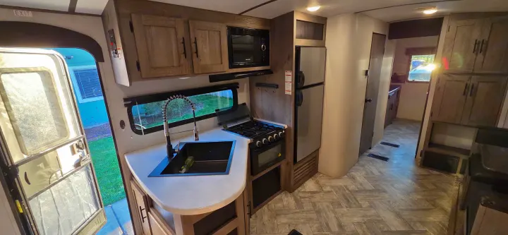 Modern rv rental kitchen area with sink, stove, microwave, and fridge. Bright, wood-toned interior, open door view.. Keystone RV Springdale 2021