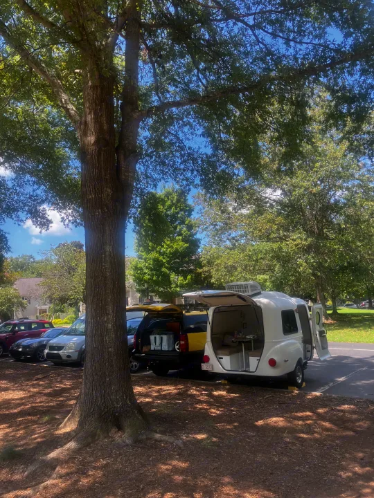 White teardrop RV rental parked under a large tree with its doors open, showing its interior, next to a yellow SUV.. Happier Camper HC1 2022